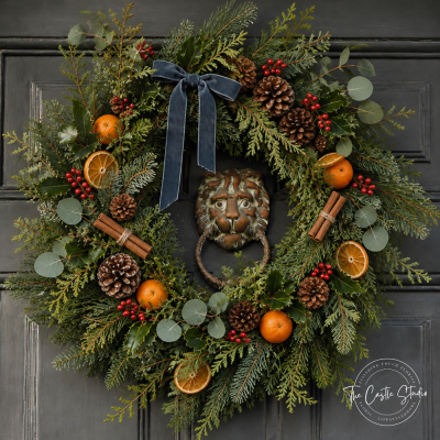The Castle Christmas Wreath - Nothing says "welcome home" quite like a wreath that commands the front door — and ours does it with quiet, old-world grandeur. The Castle Christmas Wreath is generously handcrafted from an abundant blend of pine, fir and silvery eucalyptus, giving it a full, deeply textured look that fills the doorway with the richness of the season.

Nature's finest details are layered throughout: clusters of bright red berries, a mix of natural pinecones, warm dried orange slices, and bundles of cinnamon sticks tied with twine — a subtle nod to the cosy scent of the holidays. A deep slate-blue velvet bow crowns the design, its long tails trailing elegantly downward.

Perfectly at home against panelled doors, stone walls, or a grand entrance, this is a wreath made for the home that loves a little drama with its tradition.